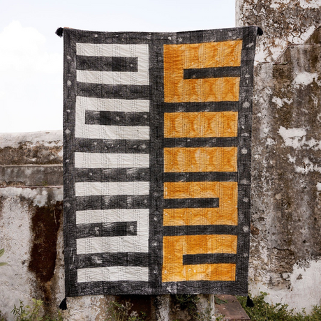 Textile art piece with geometric patterns hanging against a textured wall.