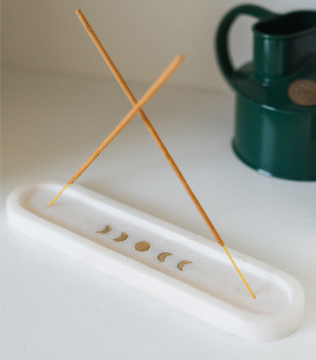 Incense stick holder with incense sticks on a white surface, with a green container in the background.