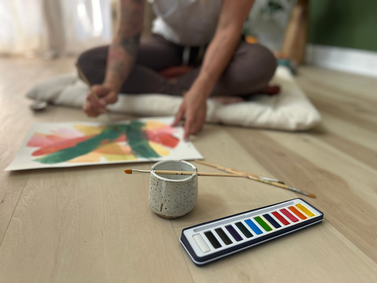 Person sitting on the floor with a colorful painting, paintbrushes, and a color swatch book.