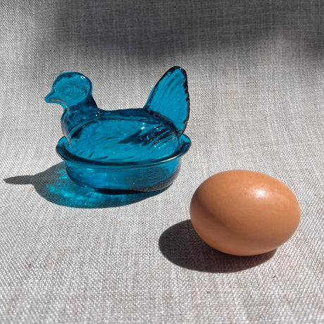 Blue nesting  hen-shaped bowl with a brown egg on a textured fabric background