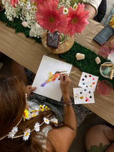 Person painting a colorful design on paper with flowers and art supplies on a table.