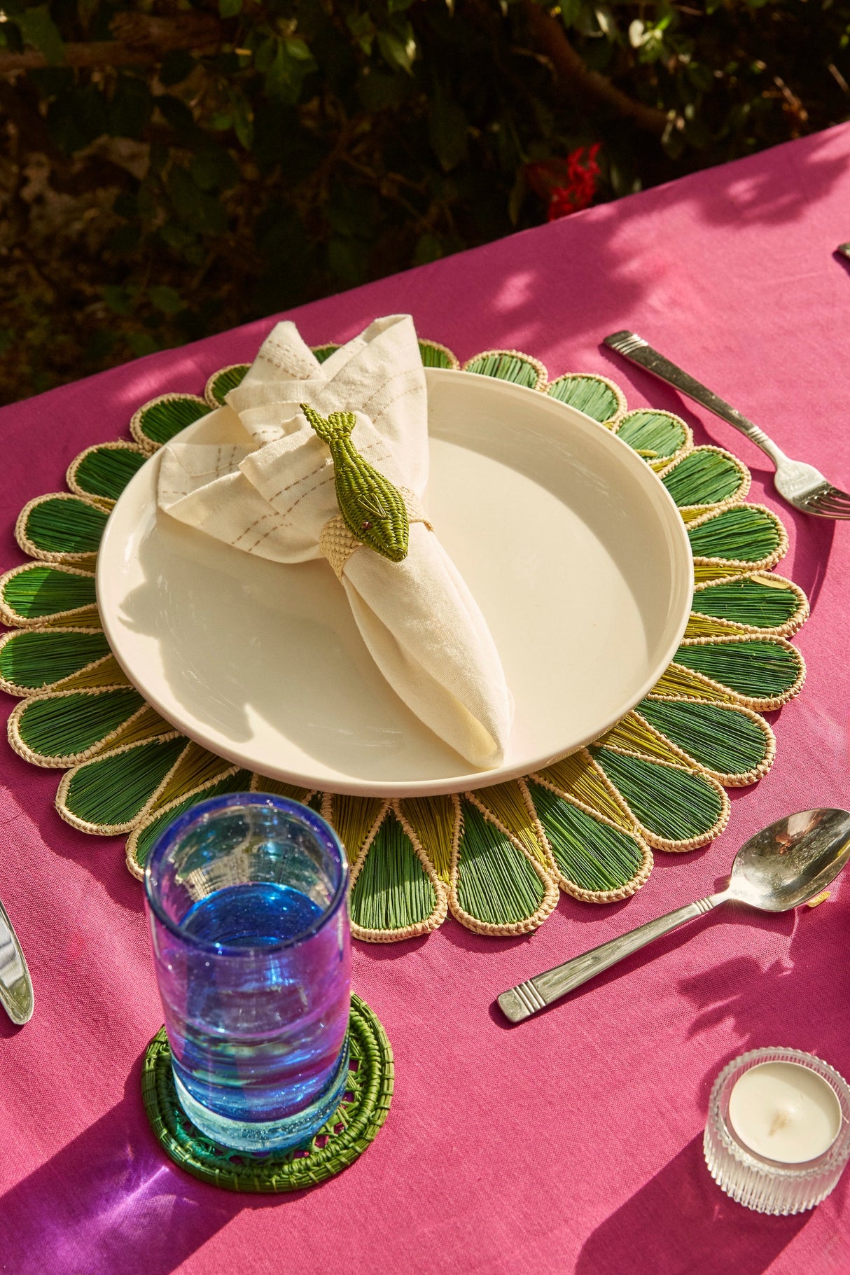 Table setting with a green placemat, white plate, folded napkin, glass of water, and candle on a pink tablecloth.