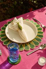 Table setting with a green placemat, white plate, folded napkin, glass of water, and candle on a pink tablecloth.