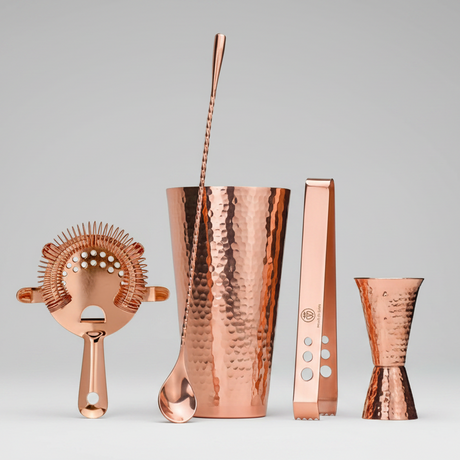 Set of copper cocktail tools including a shaker, strainer, and jigger on a light gray background.