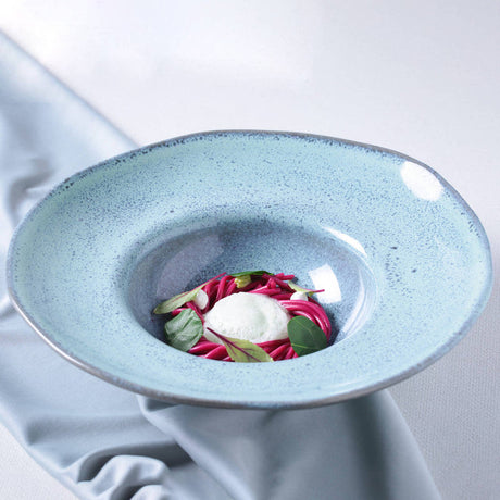 Blue ceramic bowl with a dish of pink noodles and green leaves on a white background