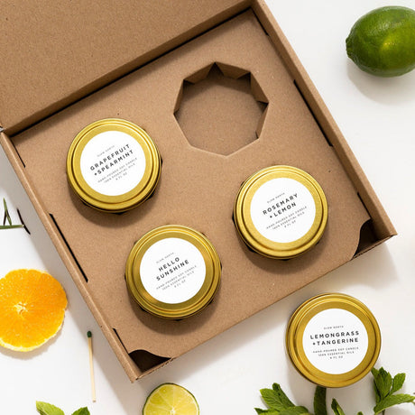 Set of four small jars with gold lids and white labels in a cardboard box, surrounded by citrus fruits and leaves.
