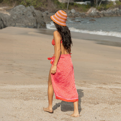 Woman in a red dress and striped hat walking on a sandy beach.