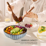 Person serving salad from a large bowl with wooden spoons, surrounded by drinks on a white table.