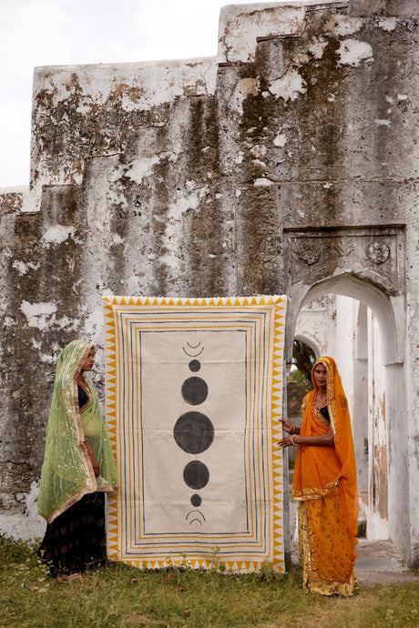 Two women holding a tapestry with celestial design in front of stone ruins