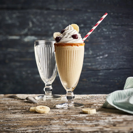 The La Rochere Antillaise Ice Cream Cup, filled with a milkshake topped with whipped cream, cherries, and banana slices, evokes classic designs while another cup sits empty on the rustic table nearby.