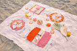 Set table with colorful plates, cutlery, and glasses on a white blanket on sand.