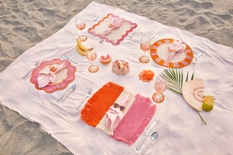 Set table with colorful plates, cutlery, and glasses on a white blanket on sand.