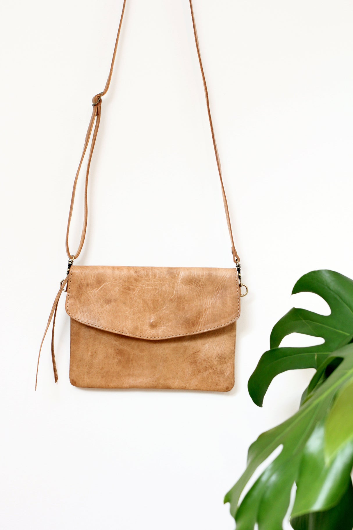 A brown full grain leather crossbody clutch with a removable strap, displayed against a white background with a green plant to the side.