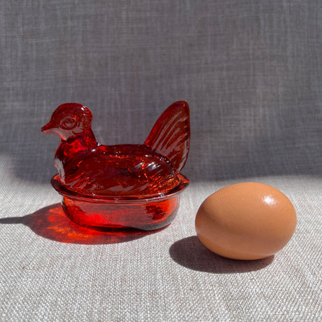 Orange glass chicken-shaped bowl next to a brown egg on a textured surface with a gray background
