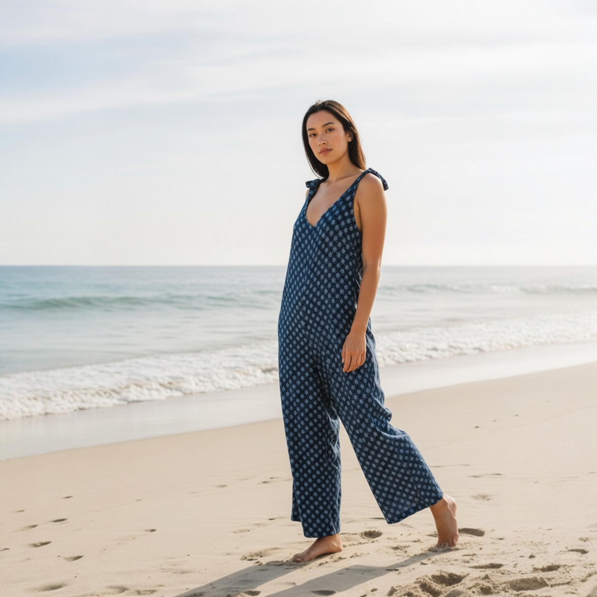 Woman in a blue patterned jumpsuit standing on a beach with ocean in the background