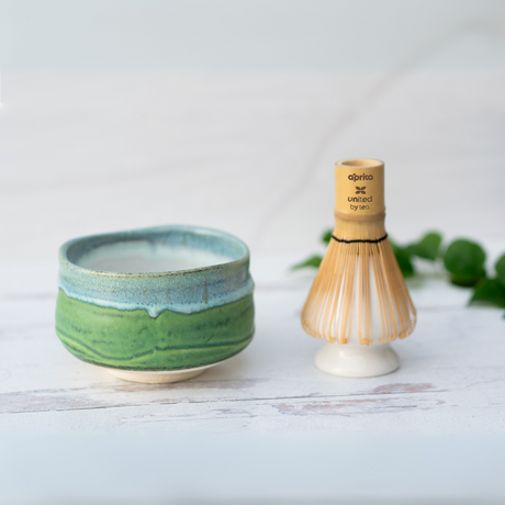 An Iguchi Deep Sea Clay Matcha Bowl, part of the Minoyaki pottery line, and a bamboo whisk sit elegantly on a white wooden surface, framed by green leaves.