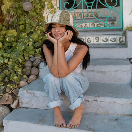 Woman sitting on steps wearing a hat and casual outfit, with greenery and a decorative wall in the background.