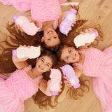Three young girls in pink dresses lying on a wooden floor holding toy bottles.
