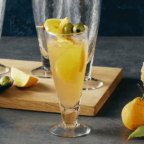 Cocktail in a tall glass with olives and lemon slices on a wooden board.