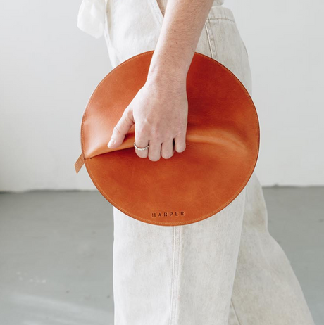 Person holding a round, brown leather clutch with 'Harper' branding against a neutral background