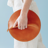 Person holding a round, brown leather clutch with 'Harper' branding against a light blue background