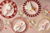 Decorative table setting with pink plates, red rimmed chargers, and floral napkins on a striped tablecloth.