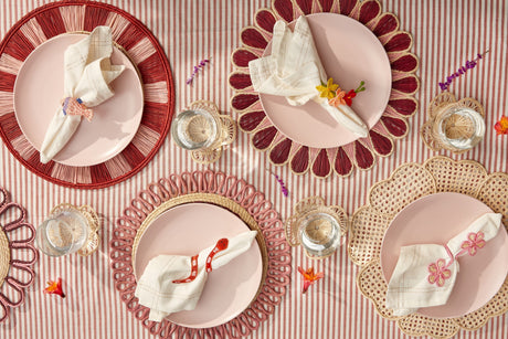 Decorative table setting with pink plates, red rimmed chargers, and floral napkins on a striped tablecloth.