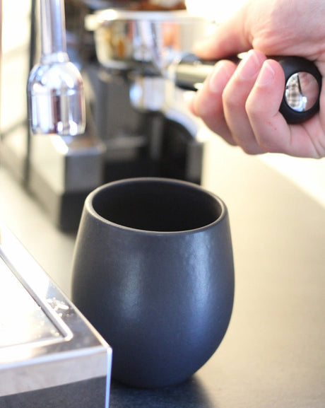Black ceramic cup on a kitchen counter with a blurred background of a coffee machine.