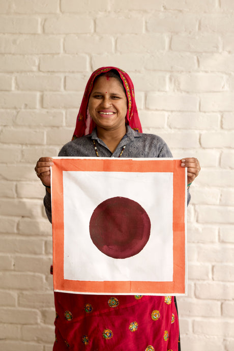 Woman holding a framed artwork with a red circle on a white background against a brick wall.