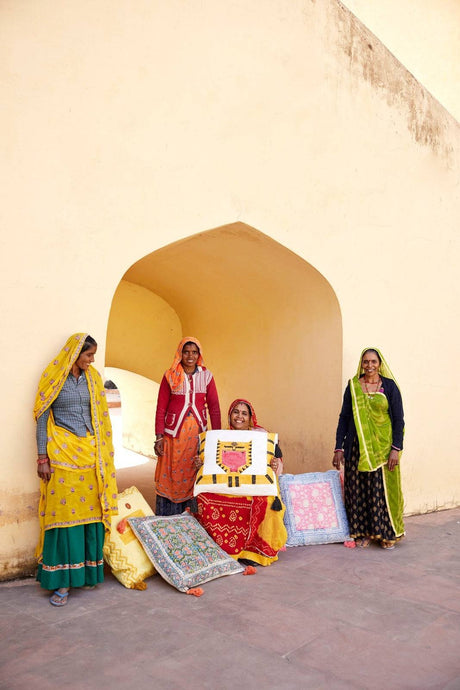 Four women in colorful traditional attire standing near a decorative table with cushions against a beige wall.