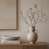 Beige vase with branches on a wooden surface against a beige wall.
