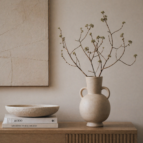 Beige vase with branches on a wooden surface against a beige wall.