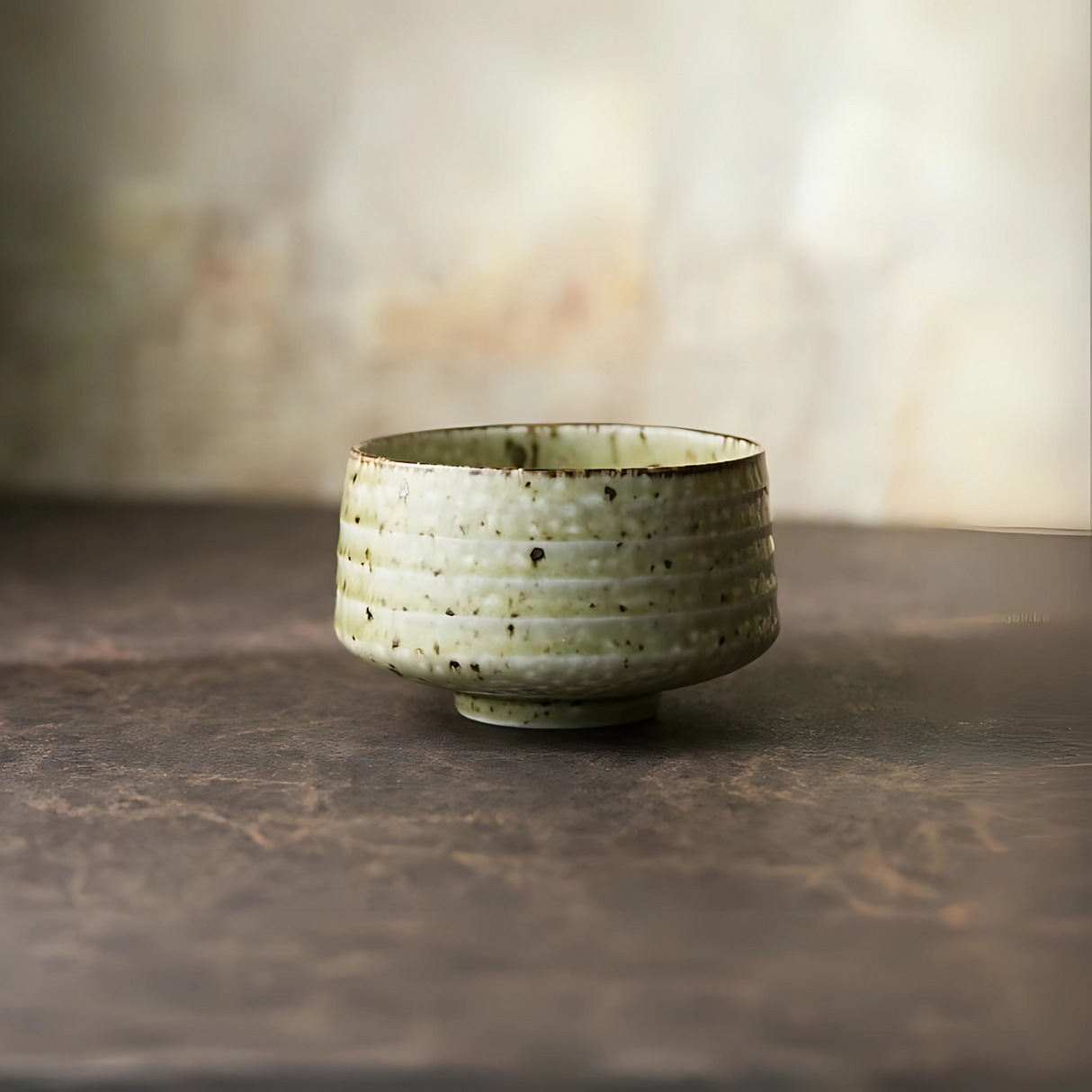 The Kaneni White Ceramic Matcha Bowl, with its speckled glaze and Japanese-inspired design, rests on a rustic surface against a softly blurred background.