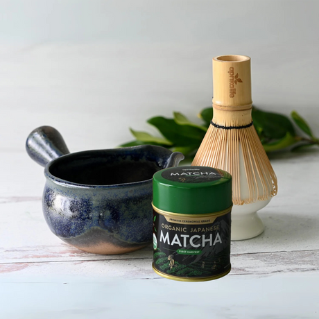 A Blue Spouted Ceramic Matcha Bowl set, including a bamboo whisk, whisk holder, and tin of organic Japanese matcha, is elegantly displayed on a light surface with scattered leaves.