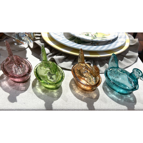 Four colorful glass hen figurines on a table setting with plates and napkins.