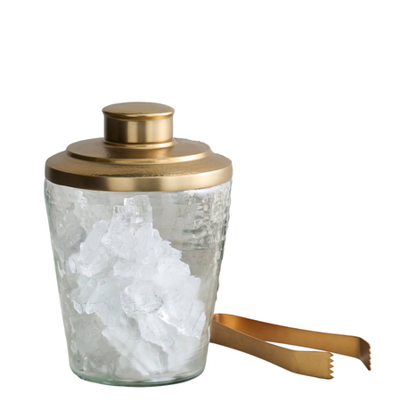 Glass cocktail shaker with gold lid and ice, surrounded by bar tools on a white background