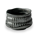 The Black Ceramic Meisen Matcha Bowl, with its textured dark finish, wavy rim, and ribbed pattern reminiscent of Minoyaki pottery, stands elegantly against a black background.