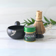 The Black Flowers Ceramic Matcha Bowl with Spout, accompanied by a tea canister, bamboo whisk, and green leaves on a light surface, is ideal for honoring the Japanese tea ceremony or as a thoughtful housewarming gift.