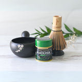 The Black Flowers Ceramic Matcha Bowl with Spout, accompanied by a tea canister, bamboo whisk, and green leaves on a light surface, is ideal for honoring the Japanese tea ceremony or as a thoughtful housewarming gift.