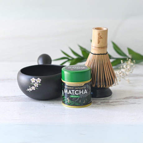 The Black Flowers Ceramic Matcha Bowl with Spout, accompanied by a tea canister, bamboo whisk, and green leaves on a light surface, is ideal for honoring the Japanese tea ceremony or as a thoughtful housewarming gift.