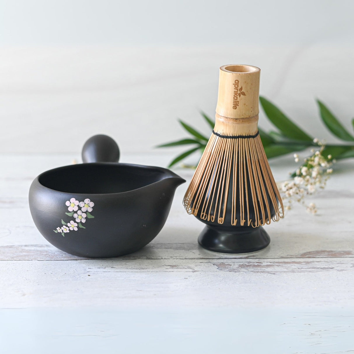 The Black Flowers Ceramic Matcha Bowl with Spout, paired with a bamboo whisk on a light wooden surface and surrounded by greenery, is perfect for Japanese tea ceremonies and makes an ideal housewarming gift for matcha enthusiasts.