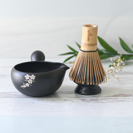 The Black Flowers Ceramic Matcha Bowl with Spout, paired with a bamboo whisk on a light wooden surface and surrounded by greenery, is perfect for Japanese tea ceremonies and makes an ideal housewarming gift for matcha enthusiasts.