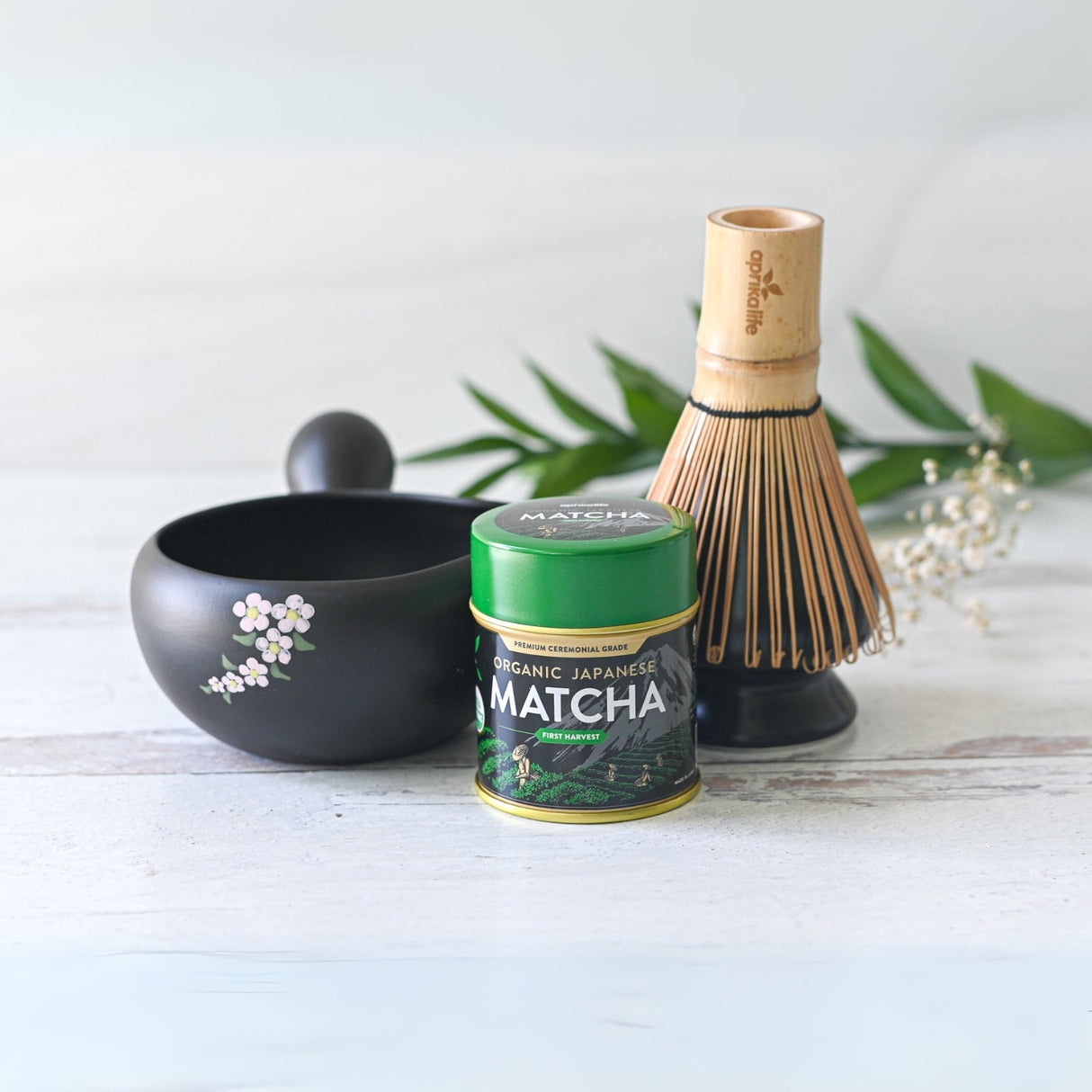 The Black Flowers Ceramic Matcha Bowl with Spout, accompanied by a tea canister, bamboo whisk, and green leaves on a light surface, is ideal for honoring the Japanese tea ceremony or as a thoughtful housewarming gift.