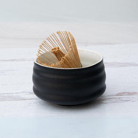 A Black and White Ceramic Matcha Bowl, crafted in Minoyaki pottery style, holds a bamboo matcha whisk on a light wooden surface.