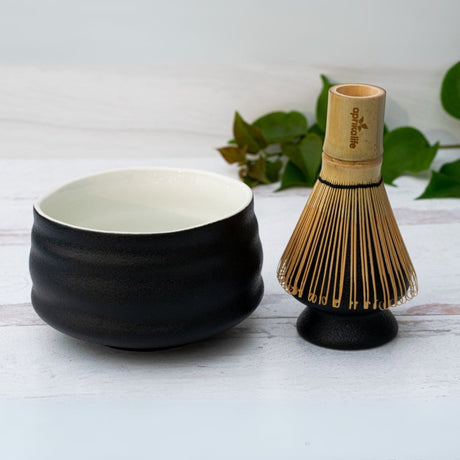 A stunning Black and White Ceramic Matcha Bowl paired with a bamboo matcha whisk rests elegantly on a white surface, framed by vibrant green leaves.