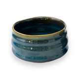 A Blue Ceramic Matcha Bowl, with a textured surface and uneven rim, sits elegantly on a black backdrop, inviting the bamboo matcha whisk to complete the timeless tea ritual.