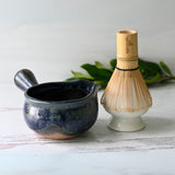 A Blue Spouted Ceramic Matcha Bowl paired with a Bamboo Matcha Whisk rests on a light surface, complemented by green leaves in the background.