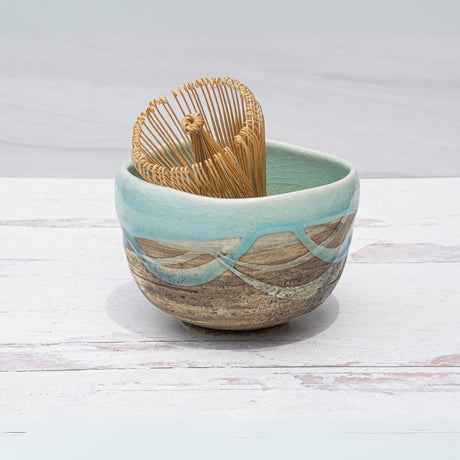 The Japanese Matcha Tea Bowl, featuring a blue and brown glaze, displays traditional Minoyaki pottery techniques while holding a bamboo whisk on a light wooden surface.