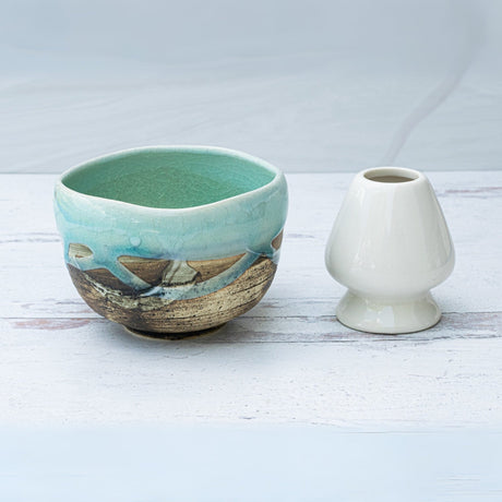 The Japanese Matcha Tea Bowl Blue & Brown Glaze, made with Minoyaki techniques, sits in turquoise and brown beside a small white vase on a light wooden surface.