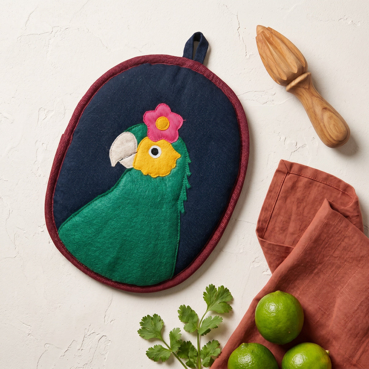 An oval fabric potholder featuring an embroidered green parrot with a pink flower on its head, set against a textured white background with a wooden citrus press, a terracotta cloth, and limes.